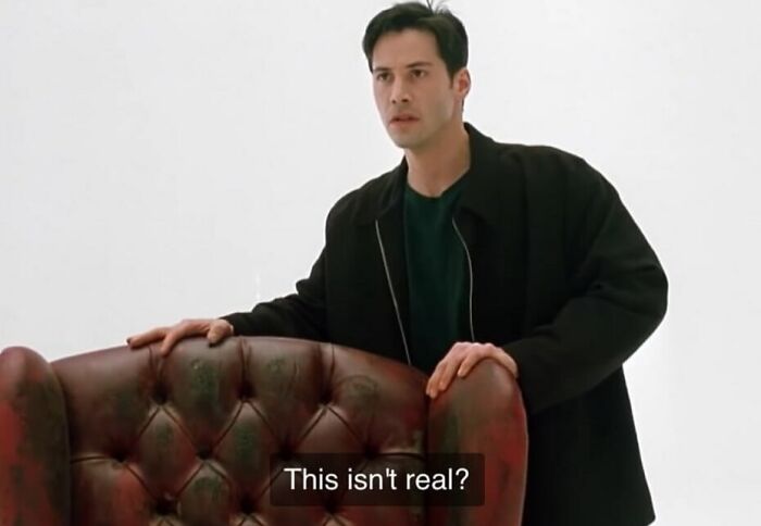Man in a black jacket by a leather armchair with the caption "This isn't real" from a nineties film scene.