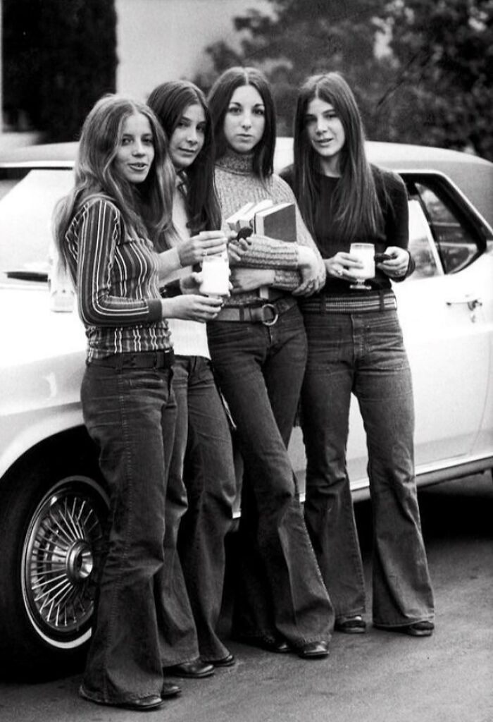 Four girls in '70s high school fashion with bell-bottoms and long hair, standing by a classic car. Four girls in '70s high school fashion with bell-bottoms and long hair, standing by a classic car.
