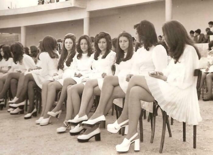High school girls in '70s fashion, wearing white dresses and platform shoes, sitting outdoors in rows of chairs. High school girls in '70s fashion, wearing white dresses and platform shoes, sitting outdoors in rows of chairs.