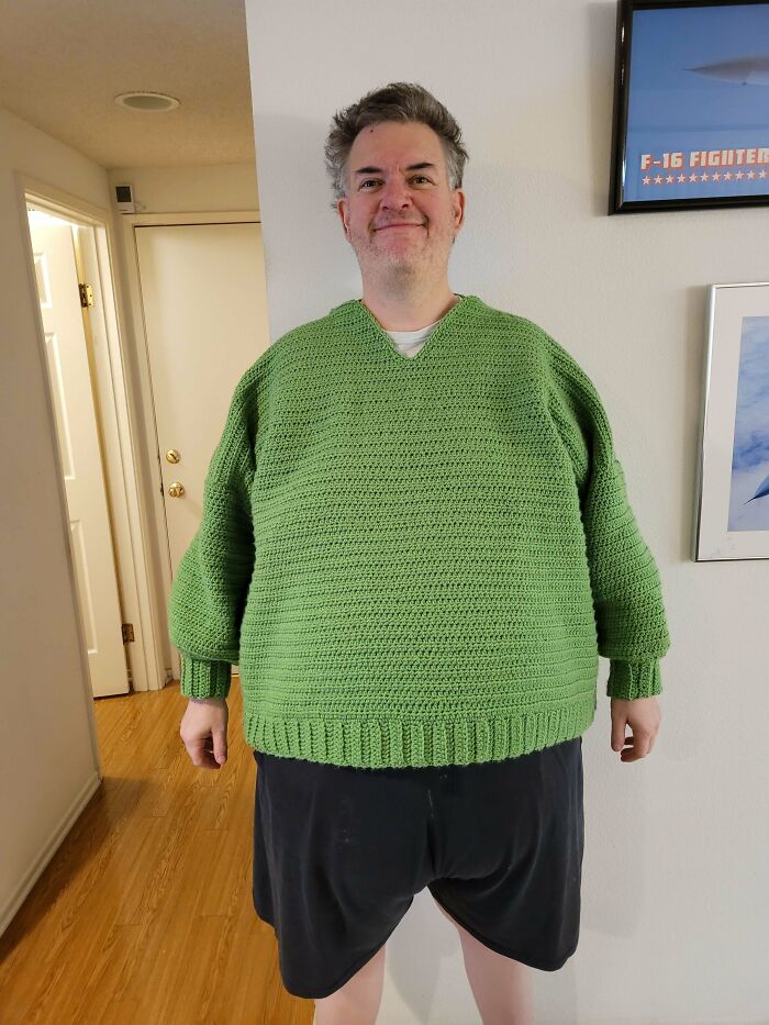 Man proudly wearing a large handmade green crochet sweater, showcasing one of his crochet projects indoors.