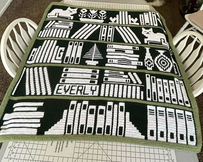 Crochet enthusiasts showcase a detailed black and white bookshelf blanket with various patterns and shapes on a table.