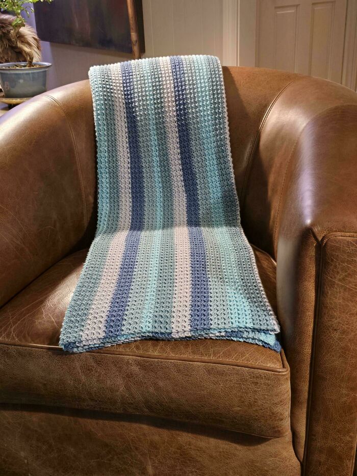 Striped blue and white crochet blanket draped over a brown leather armchair, showcasing a proud crochet project.