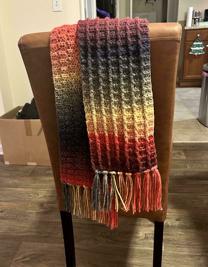 Colorful crochet scarf with fringes draped over a brown chair, showcasing textured stitch patterns and gradient yarn colors.