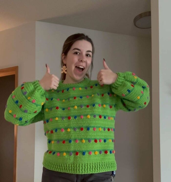 Young woman proudly showing a colorful green crochet sweater with pom poms in a home setting for crochet enthusiasts.