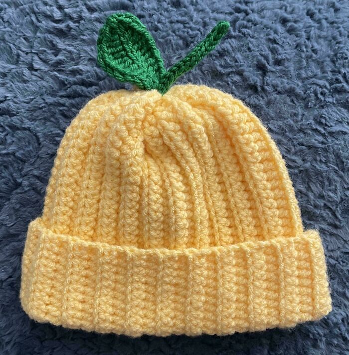 Crochet enthusiasts proudly share a yellow ribbed beanie hat with a green leaf detail on a gray textured background.