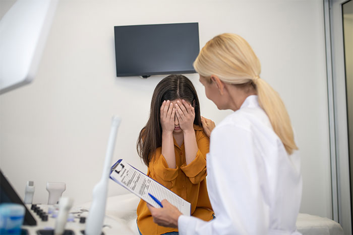 A woman in a doctor’s office, holding a clipboard, sits across from a girl covering her face, discussing daughter’s disease. A woman in a doctor’s office, holding a clipboard, sits across from a girl covering her face, discussing daughter’s disease.