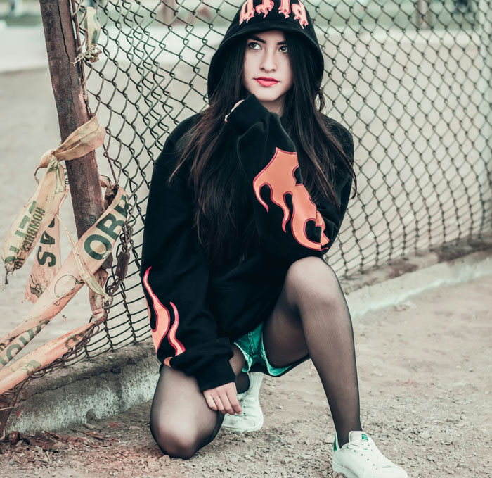 Woman in a hooded sweatshirt with flame design, kneeling by a chain-link fence, showcasing a fashion trend.