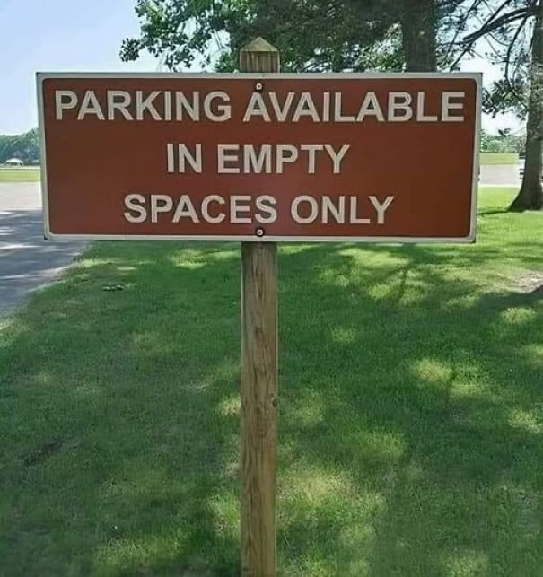 Absurd sign stating "Parking available in empty spaces only" in a park setting with grass and trees.