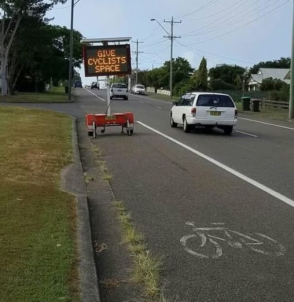 Weird sign on roadside reads "Give Cyclists Space" next to a bike lane and passing cars.