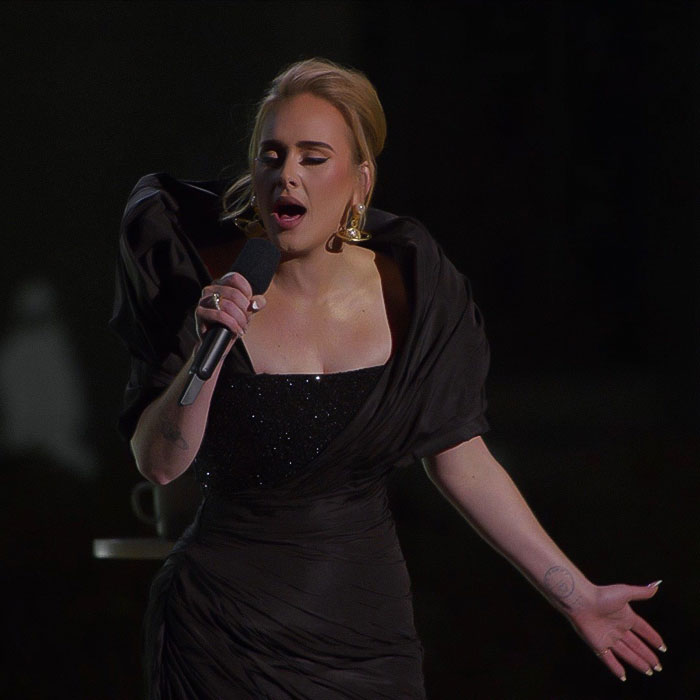 Adele performing passionately on stage in an elegant black gown, holding a microphone. Adele performing passionately on stage in an elegant black gown, holding a microphone.