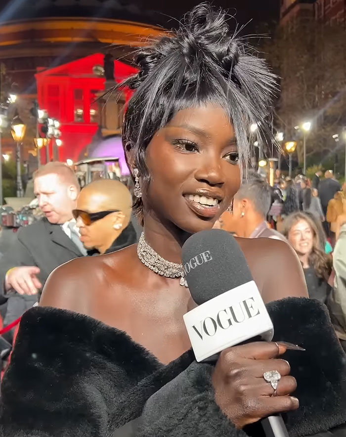 Anok Yai, a black contestant speaking at a British Fashion Awards, holding a Vogue microphone, wearing elegant jewelry and a fur stole. Anok Yai, a black contestant speaking at a British Fashion Awards, holding a Vogue microphone, wearing elegant jewelry and a fur stole.