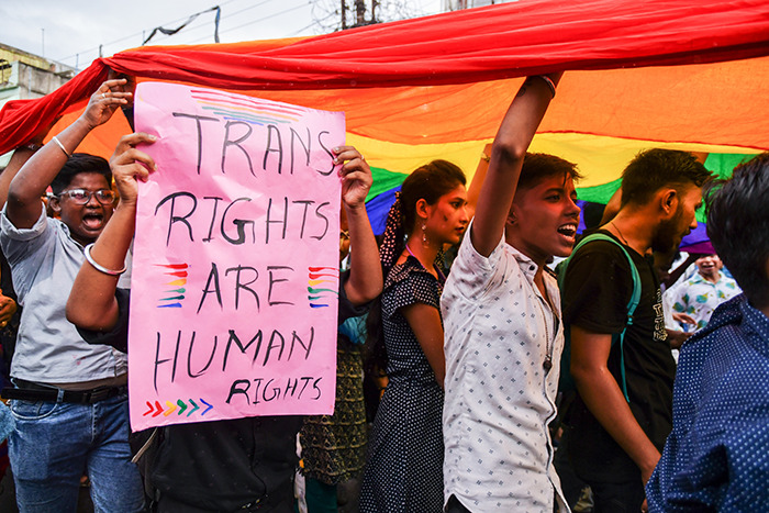 People holding a pink sign reading "Trans Rights Are Human Rights" under a rainbow flag at a protest. People holding a pink sign reading "Trans Rights Are Human Rights" under a rainbow flag at a protest.