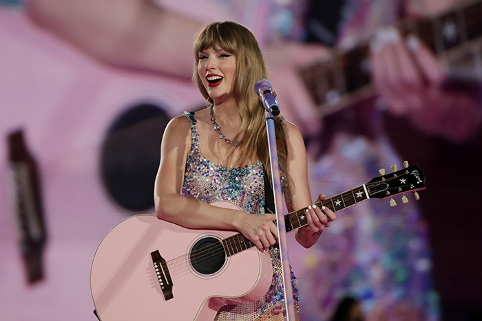 Taylor Swift smiling on stage with a pink guitar, performing live in a sparkling outfit. Taylor Swift smiling on stage with a pink guitar, performing live in a sparkling outfit.