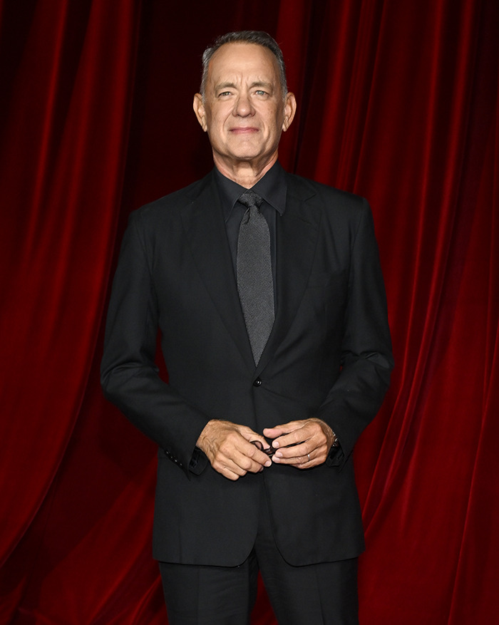 Tom Hanks in a black suit standing against a red curtain backdrop. Tom Hanks in a black suit standing against a red curtain backdrop.