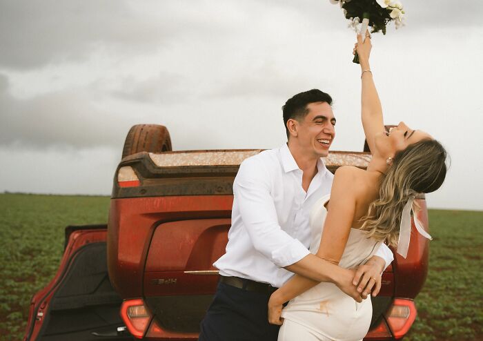 Couple laughing and posing with overturned car after accident, bride lifting bouquet joyfully in field. Couple laughing and posing with overturned car after accident, bride lifting bouquet joyfully in field.