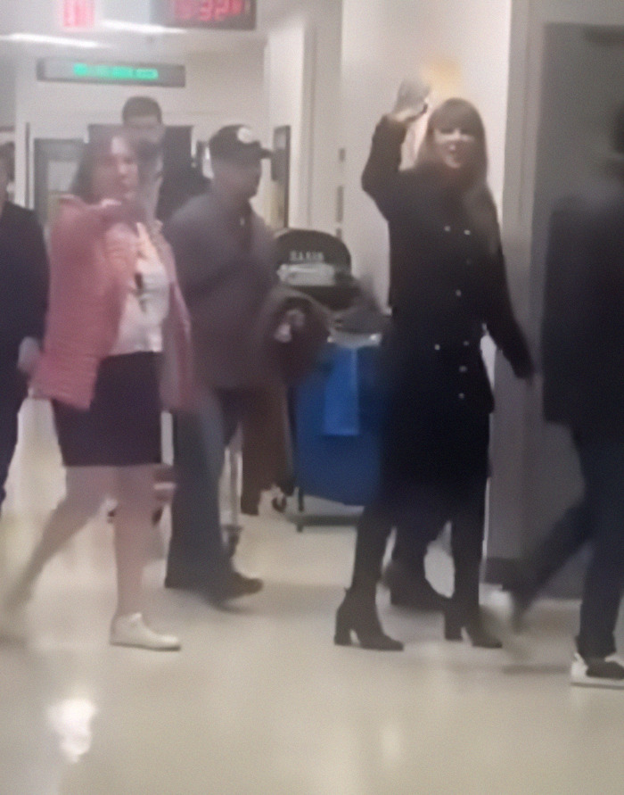 Taylor Swift in a black outfit walking through a hospital corridor, waving to hospital staff. Taylor Swift in a black outfit walking through a hospital corridor, waving to hospital staff.