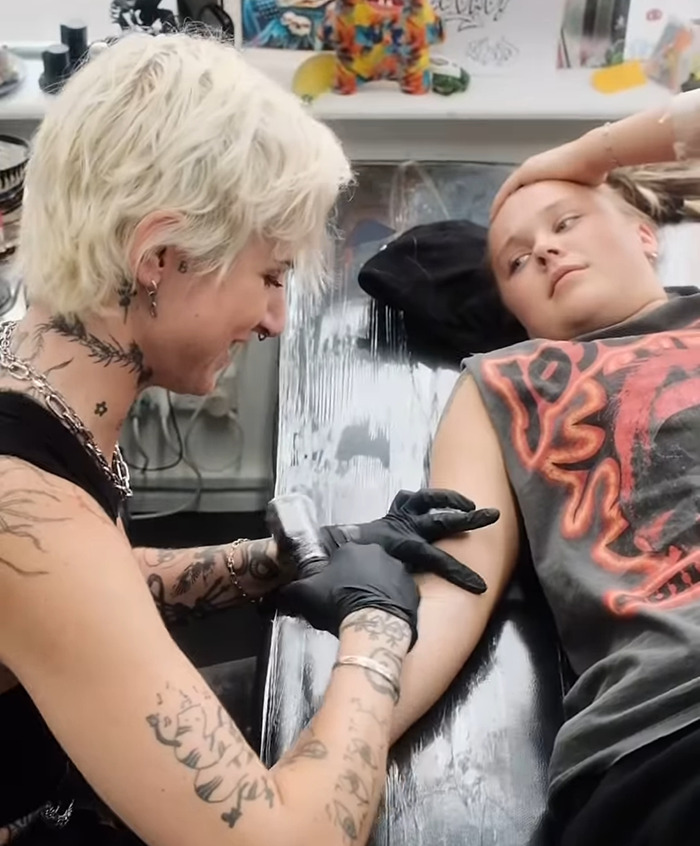 JoJo Siwa getting a new tattoo on their arm in a tattoo studio, tattoo artist with gloves and tattoos, casual setting. JoJo Siwa getting a new tattoo on their arm in a tattoo studio, tattoo artist with gloves and tattoos, casual setting.