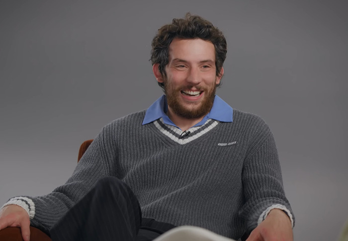 Josh O'Connor in a grey sweater and blue shirt smiles during an interview discussing Zendaya's X-rated scene. Josh O'Connor in a grey sweater and blue shirt smiles during an interview discussing Zendaya's X-rated scene.