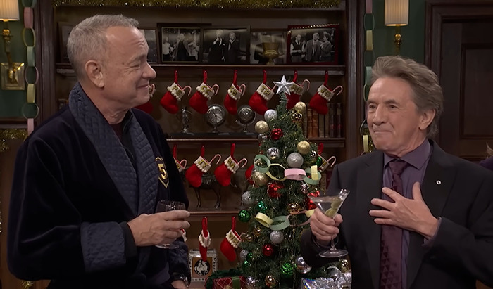 Tom Hanks on SNL, holding a drink, standing beside Martin Short near a Christmas tree. Tom Hanks on SNL, holding a drink, standing beside Martin Short near a Christmas tree.