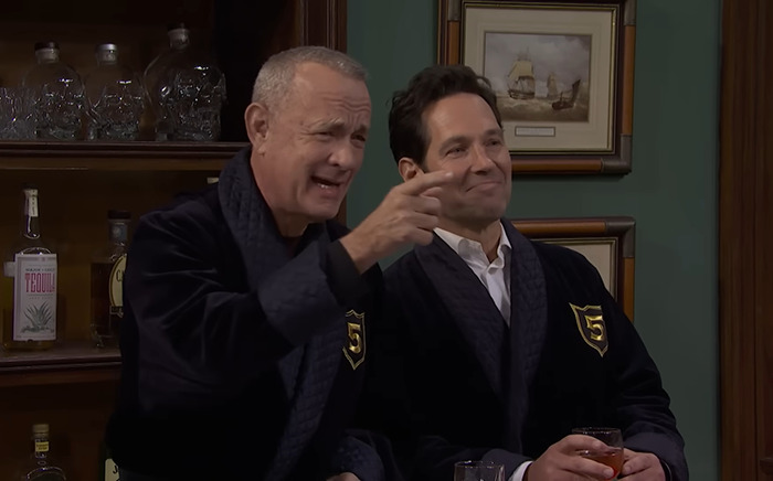 Tom Hanks and Paul Rudd in dark robes on SNL set, one gesturing with hand, holding a drink. Tom Hanks and Paul Rudd in dark robes on SNL set, one gesturing with hand, holding a drink.