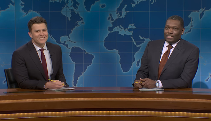 Colin Jost and co-host Michael Che at a news desk, smiling, with a world map backdrop. Colin Jost and co-host Michael Che at a news desk, smiling, with a world map backdrop.