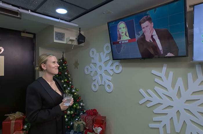 Scarlett Johansson smiles while watching Colin Jost on TV, with snowflake decorations and gifts in the background. Scarlett Johansson smiles while watching Colin Jost on TV, with snowflake decorations and gifts in the background.