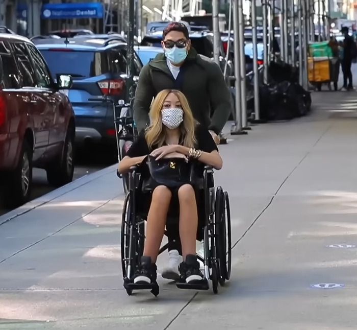 Wendy Williams in a wheelchair being pushed on a city sidewalk, both wearing masks. Wendy Williams in a wheelchair being pushed on a city sidewalk, both wearing masks.