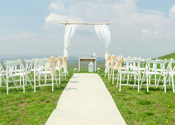 Outdoor wedding setup with white chairs and an aisle, featuring an arch adorned with sheer fabric, related to bridal event planning. Outdoor wedding setup with white chairs and an aisle, featuring an arch adorned with sheer fabric, related to bridal event planning.