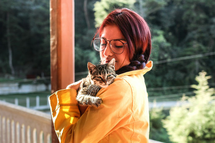 New mom holding a cat outdoors, wearing glasses and a yellow jacket, surrounded by greenery. New mom holding a cat outdoors, wearing glasses and a yellow jacket, surrounded by greenery.