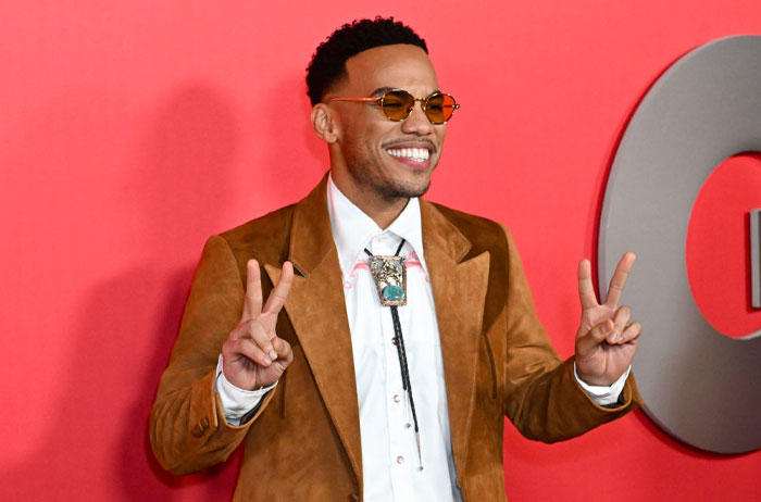 Smiling man in sunglasses at a red carpet event for Trolls 3, wearing a brown suit and bolo tie, flashing peace signs.