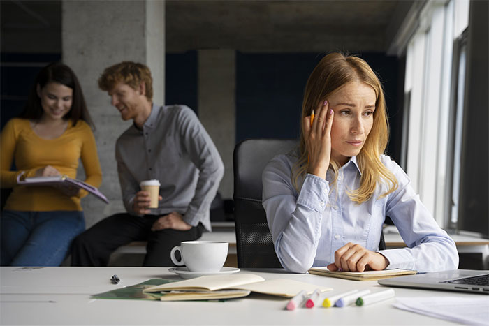 A worried woman at a desk, with a notebook and coffee, while two colleagues chat in the background. A worried woman at a desk, with a notebook and coffee, while two colleagues chat in the background.