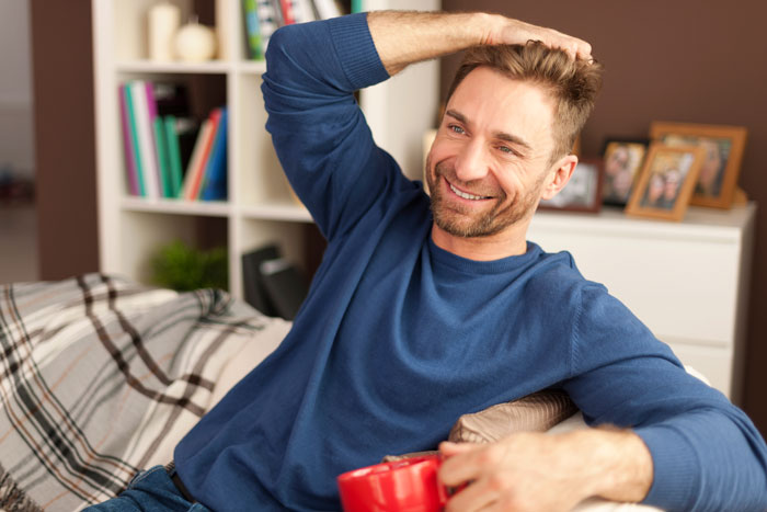 Man in blue sweater, smiling while holding a red mug, sitting on a couch. Shelves with books and photos in the background. Man in blue sweater, smiling while holding a red mug, sitting on a couch. Shelves with books and photos in the background.