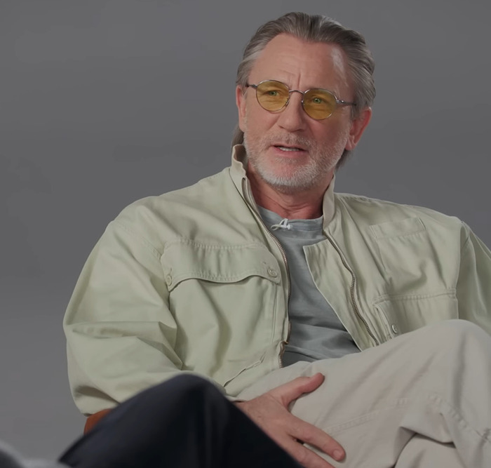 Daniel Craig in casual attire wearing yellow glasses, seated in an informal setting discussing a scene. Daniel Craig in casual attire wearing yellow glasses, seated in an informal setting discussing a scene.