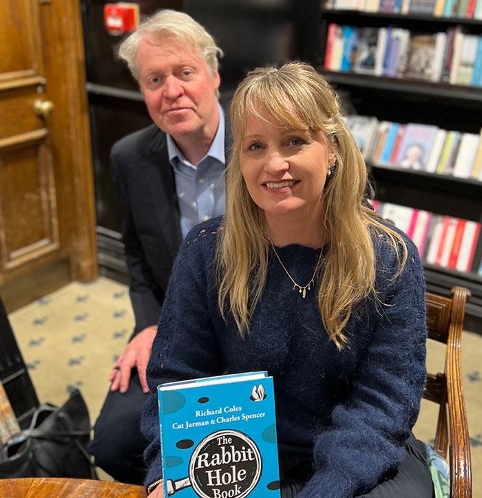 Two people in a bookstore, one holding "The Rabbit Hole Book" by Richard Coles, Cat Jarman, Charles Spencer. Two people in a bookstore, one holding "The Rabbit Hole Book" by Richard Coles, Cat Jarman, Charles Spencer.