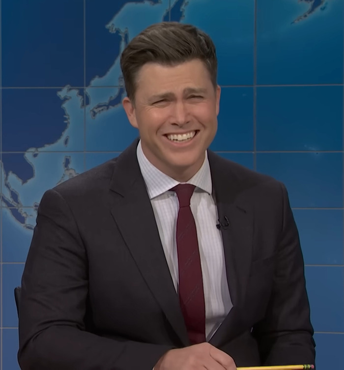Colin Jost on set, wearing a suit and smiling, reading jokes about Scarlett Johansson. Colin Jost on set, wearing a suit and smiling, reading jokes about Scarlett Johansson.