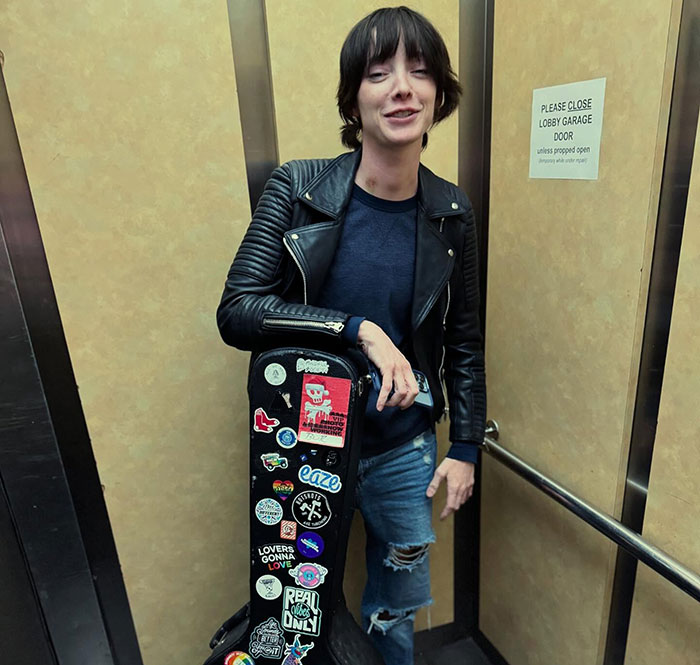 A person in an elevator wearing a leather jacket, holding a sticker-covered guitar case; related to "Oppenheimer" star. A person in an elevator wearing a leather jacket, holding a sticker-covered guitar case; related to "Oppenheimer" star.