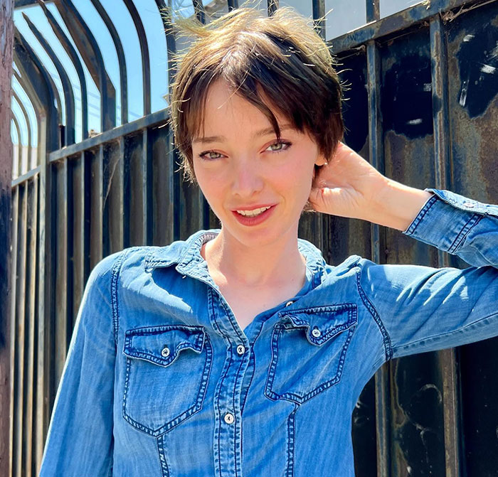 A person outdoors in denim, smiling in front of a metal fence, trans masculine non-binary theme prominent. A person outdoors in denim, smiling in front of a metal fence, trans masculine non-binary theme prominent.