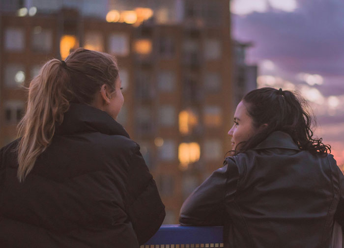 Two friends in jackets chatting on a balcony at sunset with urban buildings in the background. Two friends in jackets chatting on a balcony at sunset with urban buildings in the background.