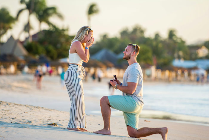 A man proposing on a beach while a woman reacts with surprise, reflecting on proposing. A man proposing on a beach while a woman reacts with surprise, reflecting on proposing.