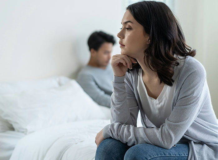 Woman deep in thought sitting on a bed, man in the background, focusing on family conflict theme. Woman deep in thought sitting on a bed, man in the background, focusing on family conflict theme.