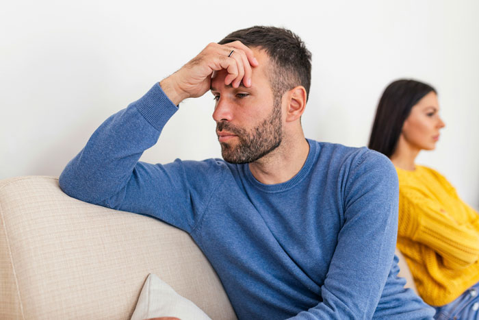Husband in blue sweater sits on couch looking upset; wife in yellow sweater sits in background, symbolizing name veto conflict. Husband in blue sweater sits on couch looking upset; wife in yellow sweater sits in background, symbolizing name veto conflict.