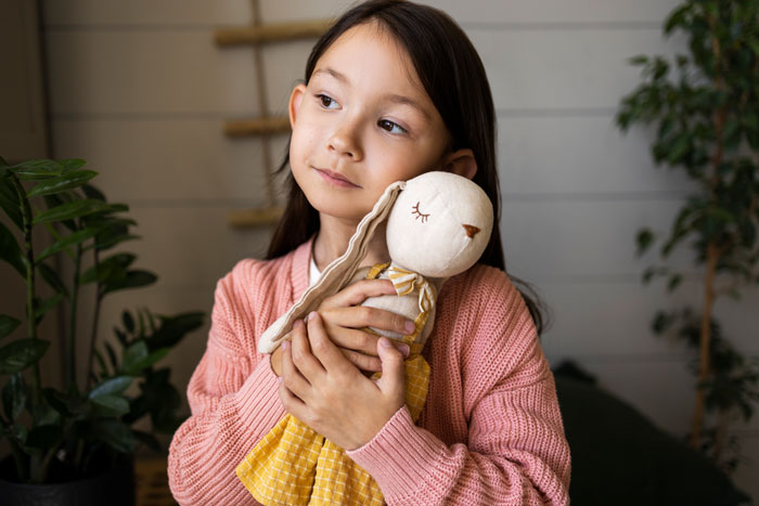 Child with Asian features holding a stuffed doll, wearing a pink sweater, looking pensive. Child with Asian features holding a stuffed doll, wearing a pink sweater, looking pensive.
