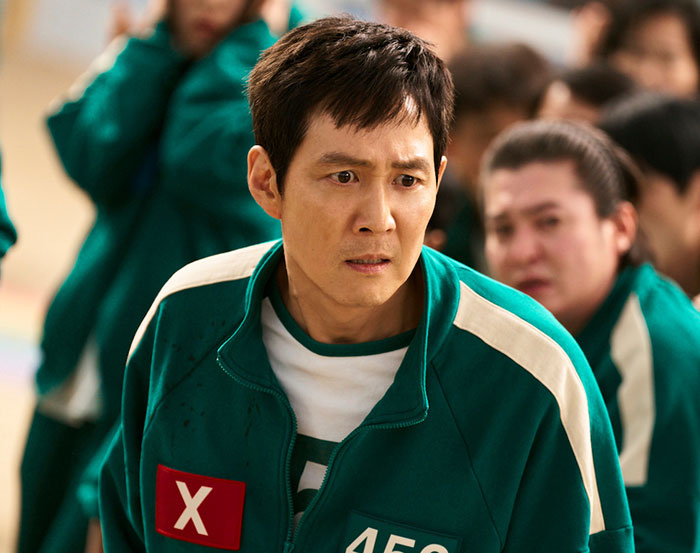 Squid Game 2 character in green tracksuit looks concerned during a tense scene. Squid Game 2 character in green tracksuit looks concerned during a tense scene.