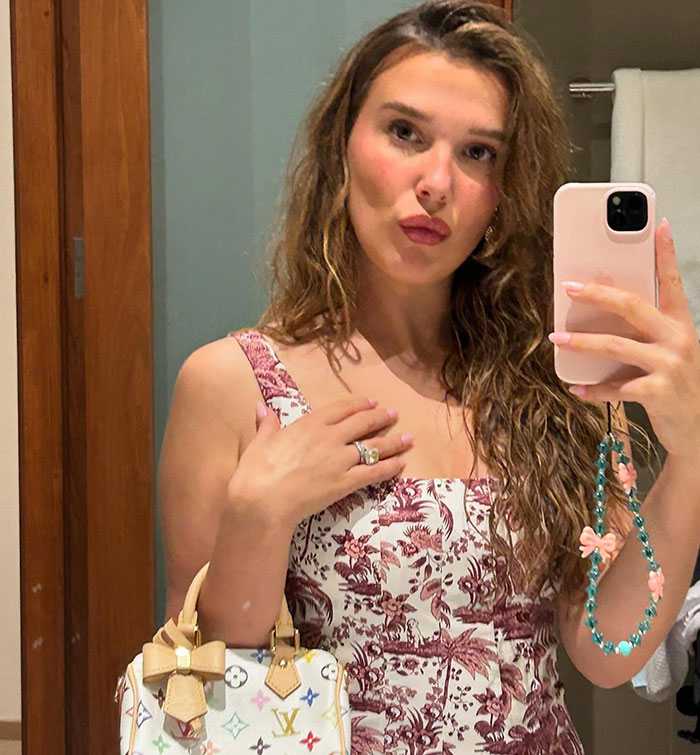 Young woman taking a mirror selfie, wearing a patterned dress and holding a designer bag, looks older. Young woman taking a mirror selfie, wearing a patterned dress and holding a designer bag, looks older.
