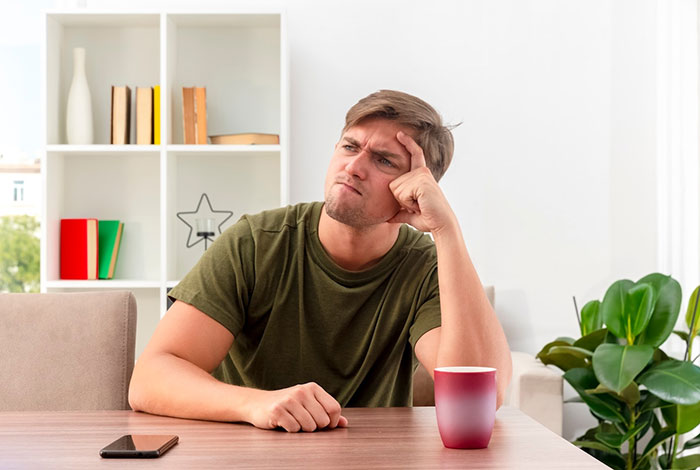 Young man in a green shirt sitting at a table, looking thoughtful and holding his forehead, phone nearby. Young man in a green shirt sitting at a table, looking thoughtful and holding his forehead, phone nearby.