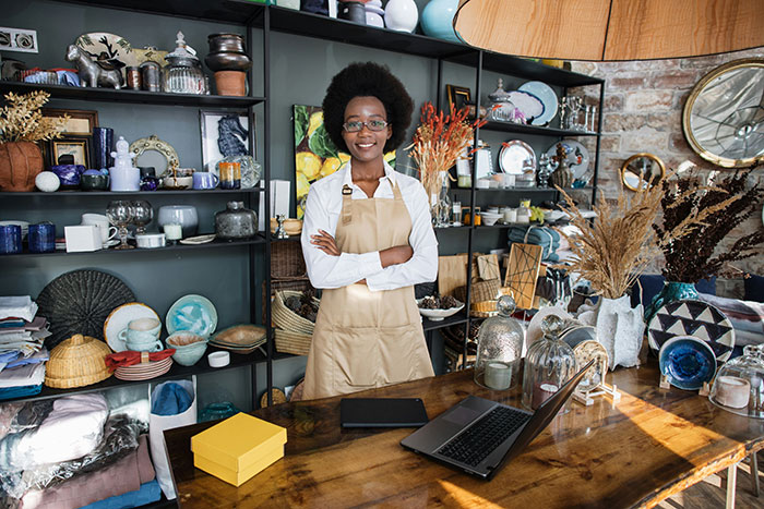 Person in a shop setting, standing confidently behind a table with a laptop, surrounded by various items, challenging stereotypes.