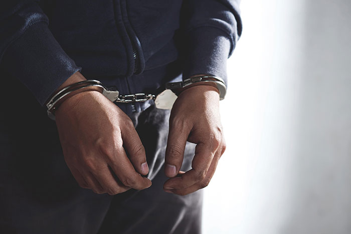 Person handcuffed in front view, wearing a dark jacket, representing a neighbor arrested after a noise complaint. Person handcuffed in front view, wearing a dark jacket, representing a neighbor arrested after a noise complaint.