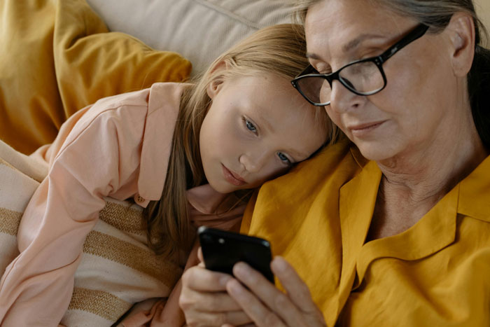Elderly woman and young girl on a couch looking at a smartphone, the woman in glasses wearing a yellow shirt. Elderly woman and young girl on a couch looking at a smartphone, the woman in glasses wearing a yellow shirt.