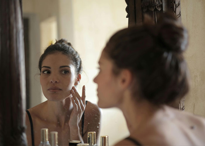 Person looking at themselves in a mirror, touching face, indoors, reflecting on healing from a rare birth defect. Person looking at themselves in a mirror, touching face, indoors, reflecting on healing from a rare birth defect.
