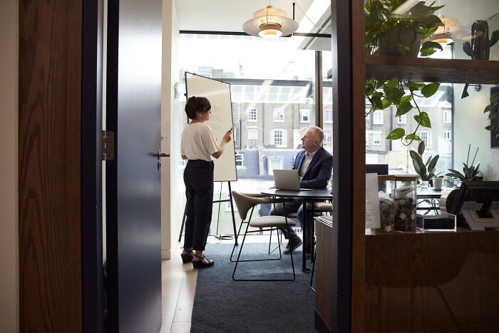 Office meeting with a woman presenting and a man seated with a laptop, highlighting changing occupations.